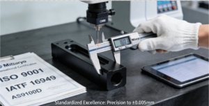 A gloved hand using a digital caliper to perform precise measurement on a black anodized aluminum prototype component, with international quality standards (ISO, IATF, AS9100) visible in the background.
